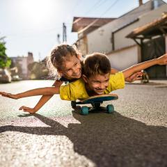A happy small boy and a girl lying on a skateboard with arms outstreched as though flying. They are on the road, there are no cars and the sun is shining.