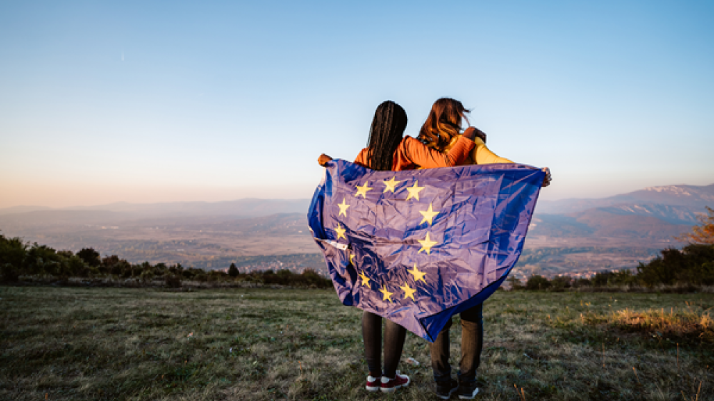 Two women standng in remote landscape with EU flag wrapped around them